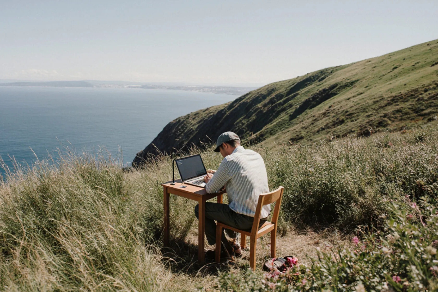 Man working on a laptop at a desk on a grassy clifftop above the sea